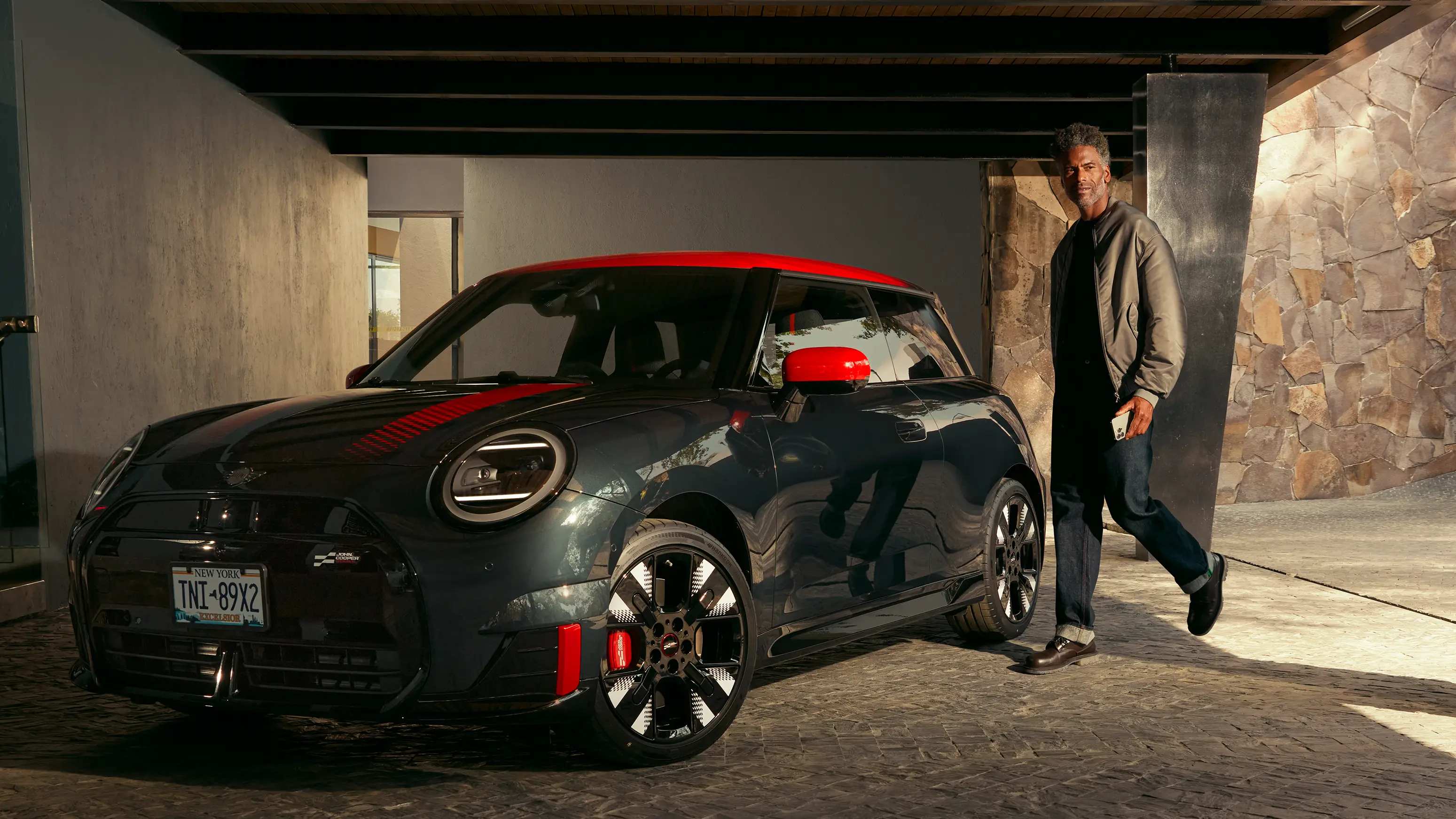 Person walking past a MINI John Cooper Works in Midnight Black II with Chili Red roof parked under a covered driveway.